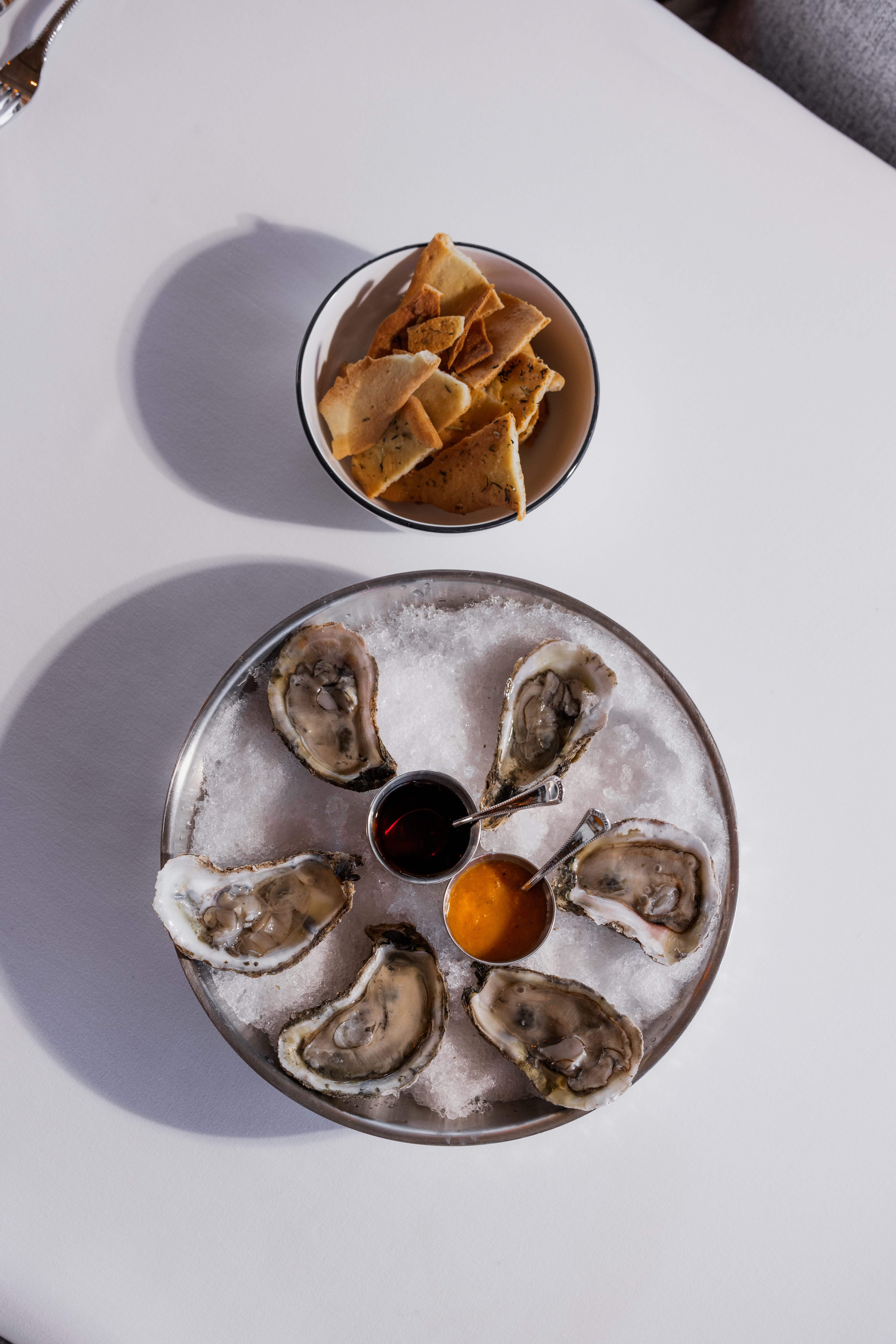 a plate of raw oysters on ice with two sauces and a bowl of crackers next to it