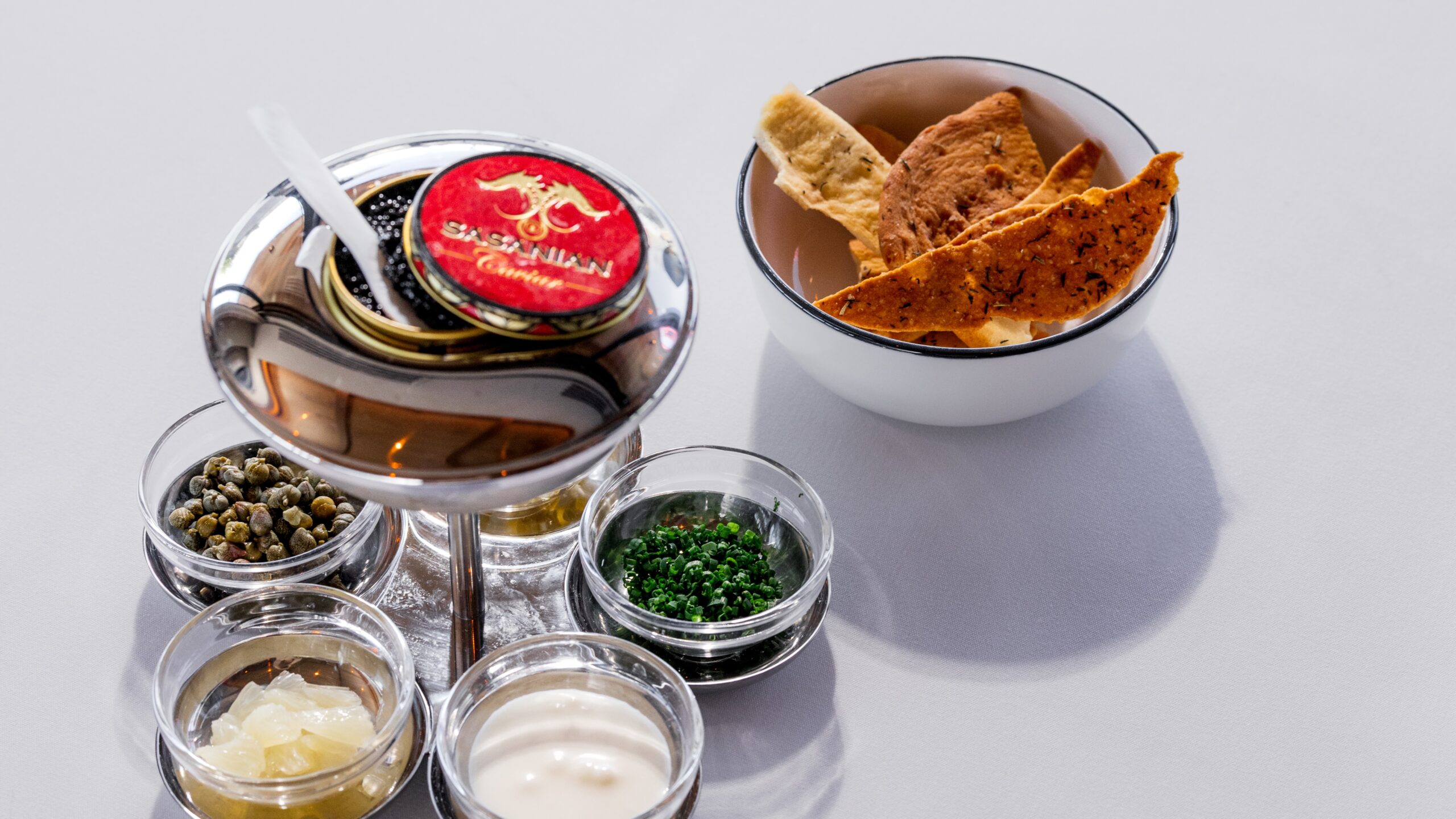 A tin of caviar on ice over bowls of chives, creme fraiche and capers with a bowl of lavash next to it
